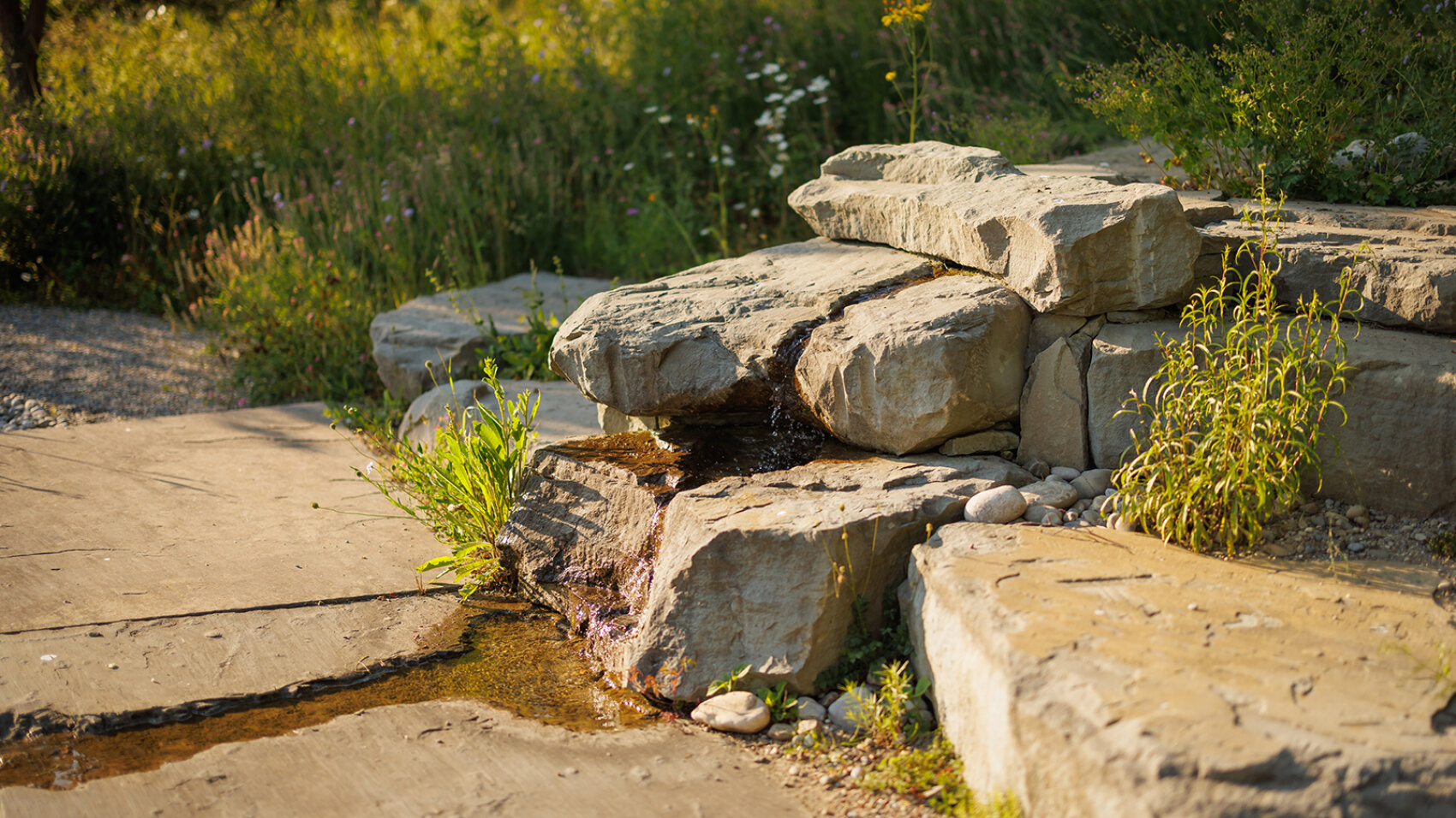 Flache Steine, die zu einem kleinen, natürlich wirkenden Springbrunnen aufgeschichtet sind, an dem das Wasser herunterrieselt, umgeben von grünem Gras und Pflanzen in einem sonnenbeschienenen Garten.