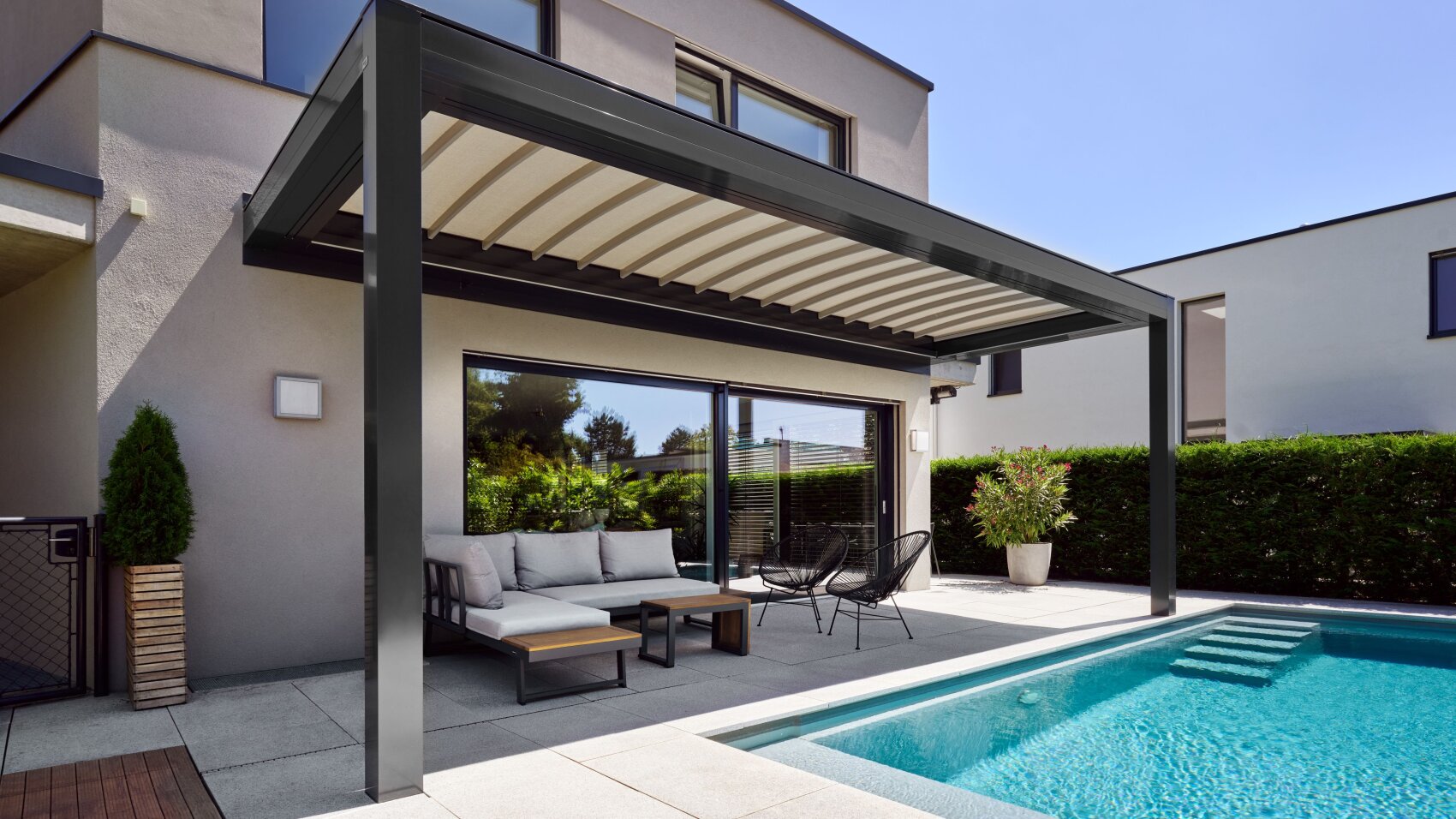 Moderne Terrasse im Hinterhof mit überdachtem Sitzbereich mit Sofa, Stühlen und Tisch neben einem Swimmingpool. Der Raum ist umgeben von Grün und modernen Hauswänden unter einem klaren blauen Himmel.