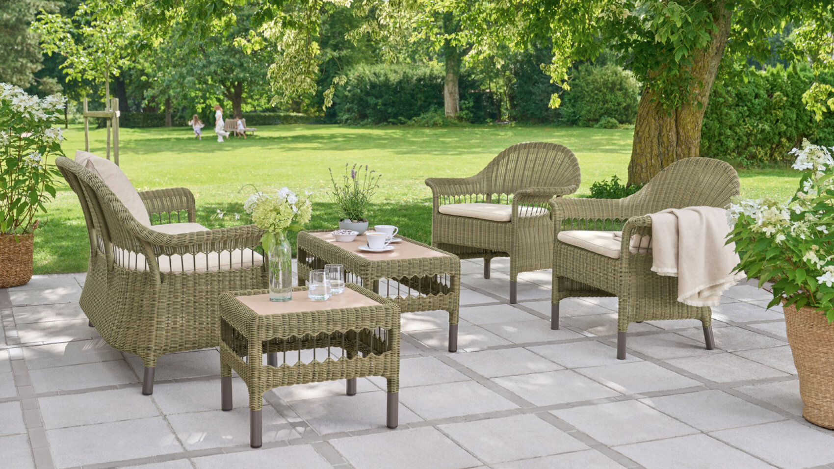 Wicker patio furniture with a sofa, two armchairs, and two tables arranged on a stone patio in a green garden. There are cups, glasses, flowers, and a throw blanket on the tables and chair. Lush trees and grass are in the background.