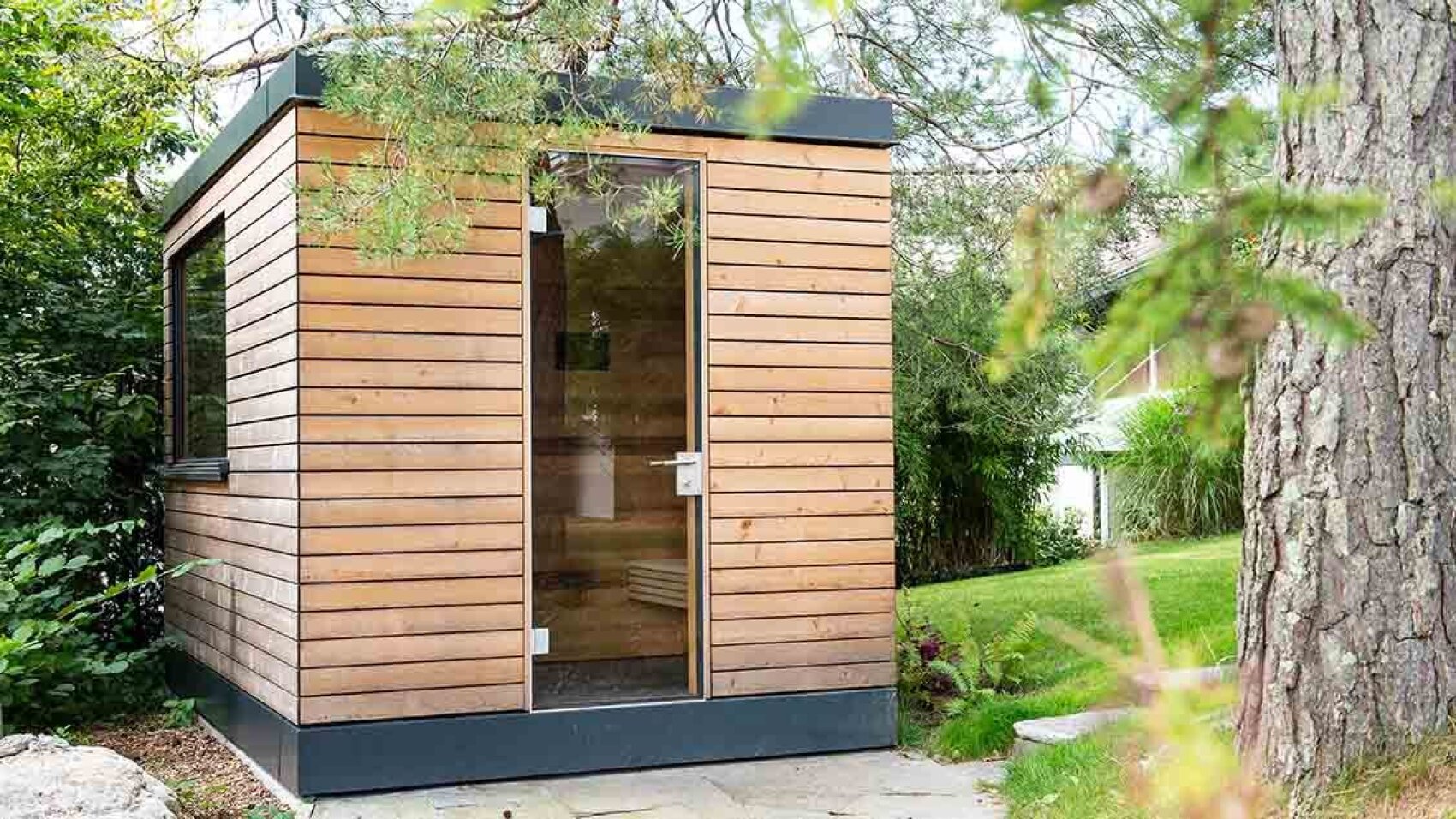 Ein kleiner, moderner Holzschuppen mit einer Glastür und horizontaler Verkleidung steht in einem üppigen, grünen Garten, umgeben von Bäumen und Pflanzen.