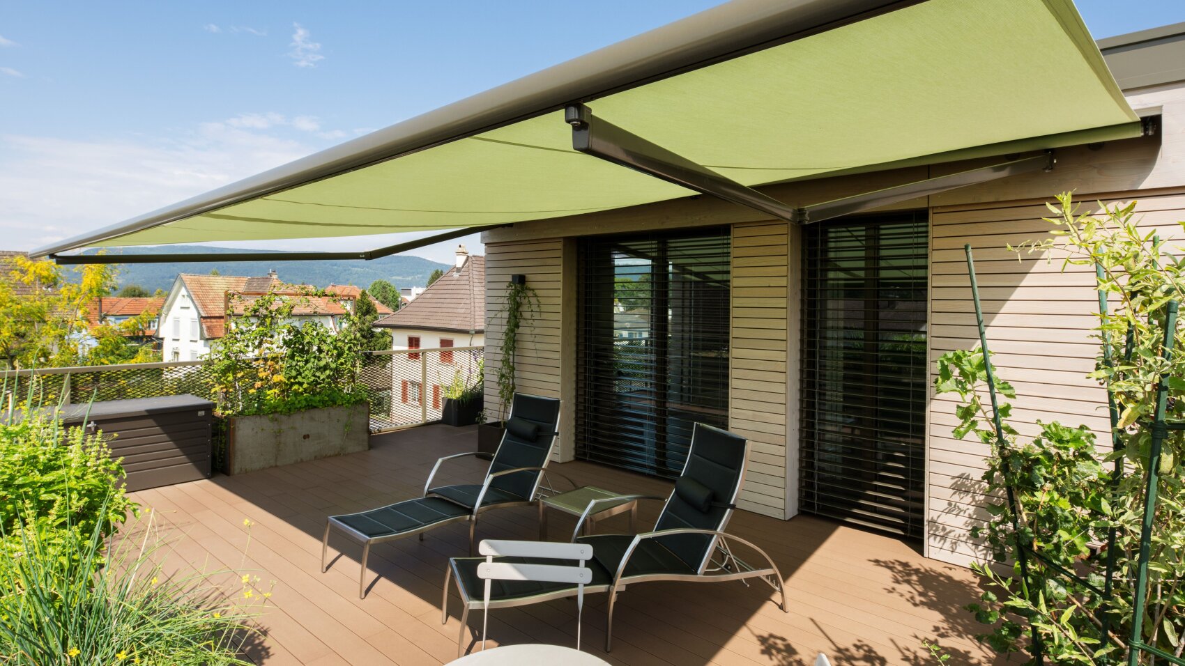 Eine moderne Terrasse mit zwei Loungesesseln und einem kleinen Tisch befindet sich unter einer großen grünen, ausfahrbaren Markise. Die Terrasse ist von Pflanzen umgeben und bietet einen Blick auf die benachbarten Häuser und die Landschaft in der Ferne.