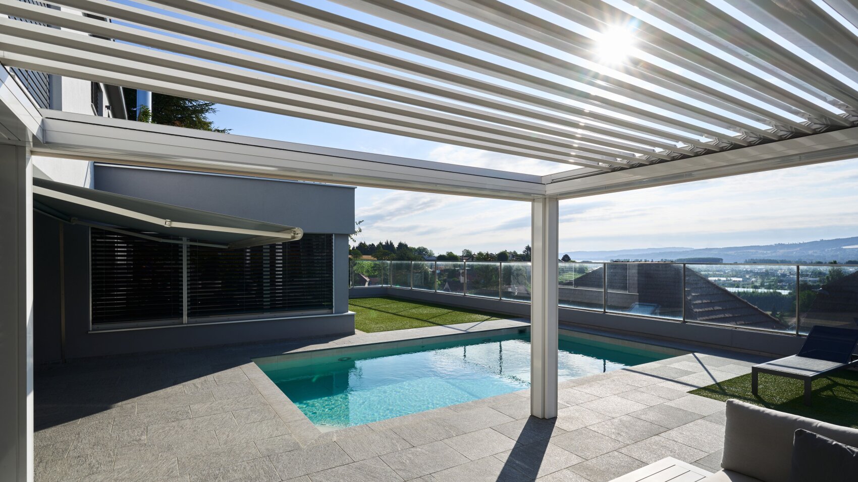 A modern patio with a small swimming pool, shaded by a white pergola with slats. Sunlight shines through the slats, creating shadows on the stone patio. There is outdoor seating and a view of hills and water in the background.