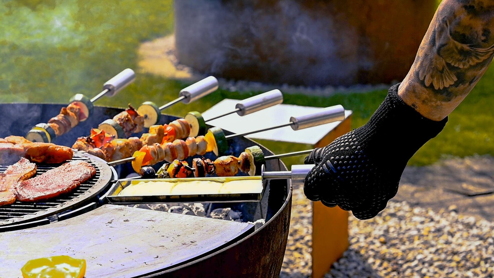 A person wearing a black glove turns skewers of grilled vegetables and meat on an outdoor grill, with smoke rising and a meadow in the background.