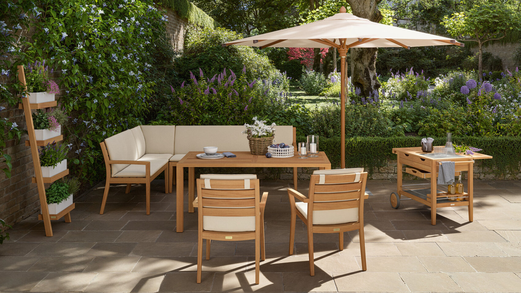 A wooden patio dining set with cushioned chairs and a parasol is arranged on a stone terrace. Nearby are a mobile serving cart, a vertical herb planter, and a lush, colorful garden in the background.