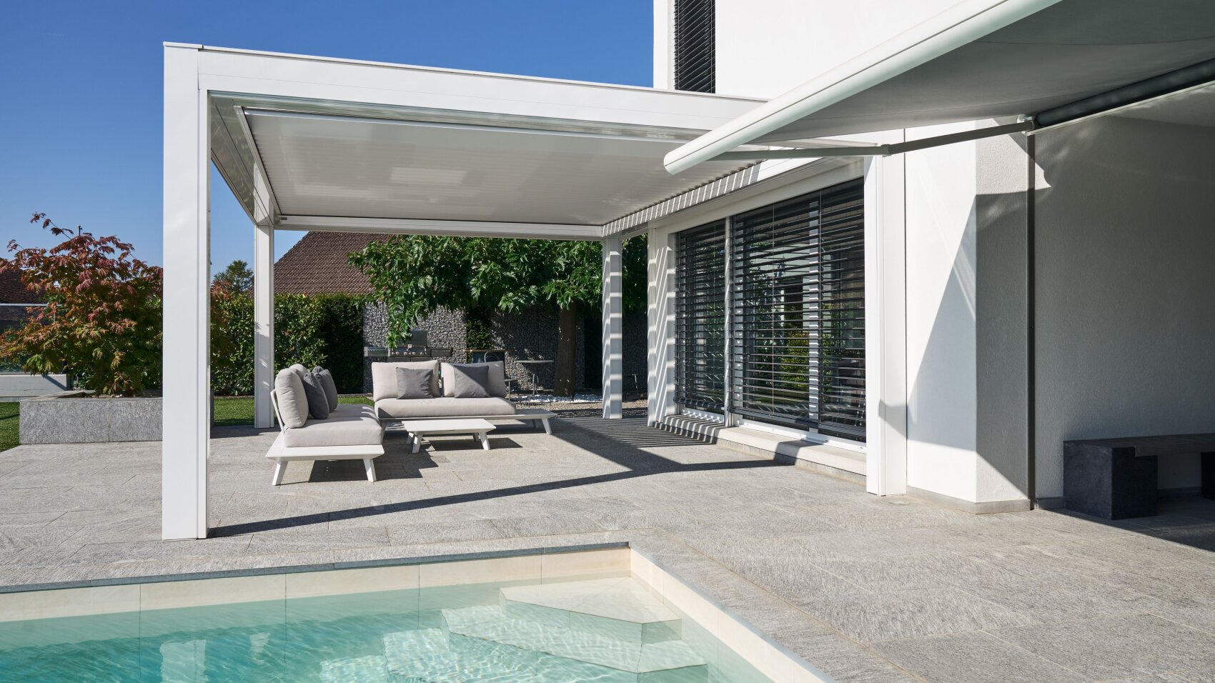 Modern patio with lounge chairs and cushions under a white pergola, next to a swimming pool. The house has large windows with blinds and an outdoor shaded seating area. Trees and greenery are visible in the background.