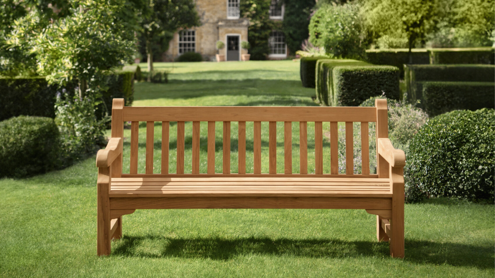 A wooden bench sits on a well-manicured lawn in a formal garden, surrounded by trimmed hedges and greenery, with a large stone house in the background.