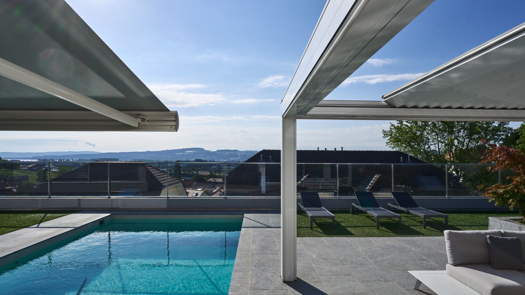 A modern outdoor pool area with lounge chairs and a partial awning, overlooking rooftops and a distant landscape under a blue sky with scattered clouds.
