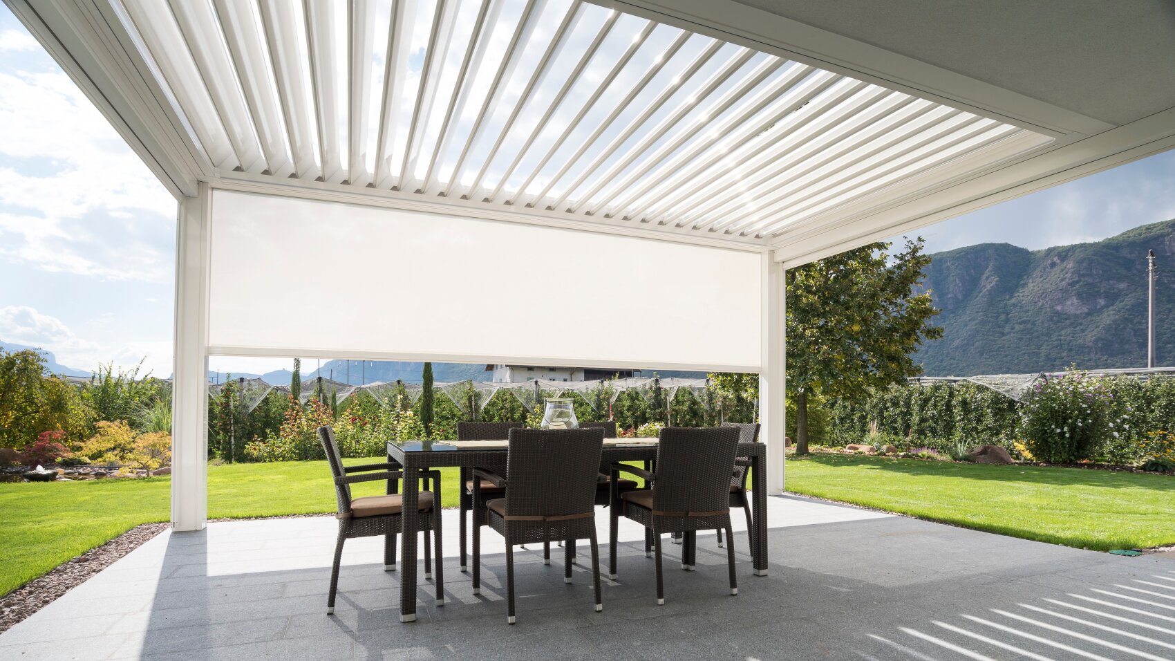 Eine moderne Außenterrasse mit einem Pergoladach aus Latten, einem rechteckigen Tisch und sechs Korbstühlen bietet an einem sonnigen Tag einen Blick auf einen Garten mit Bergen im Hintergrund.