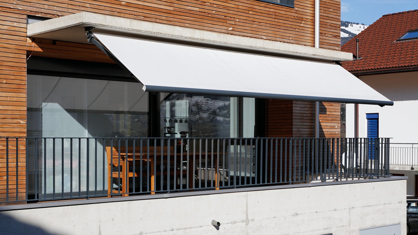 Ein moderner Balkon mit Metallgeländer, beschattet von einer großen, ausfahrbaren weißen Markise, die an einem mit Holz verkleideten Gebäude angebracht ist. Auf dem Balkon befinden sich ein Holztisch und Stühle.
