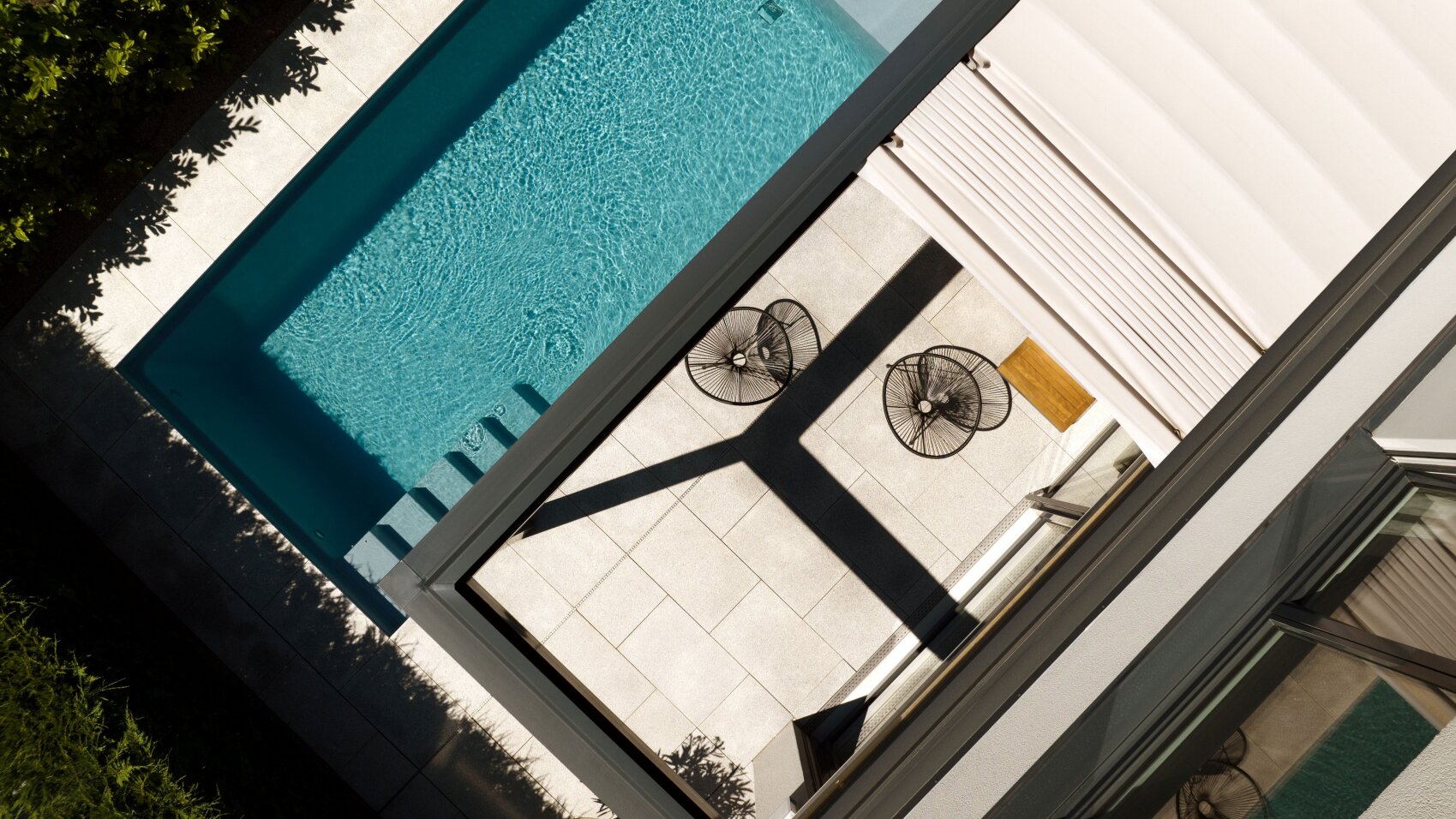 Ein moderner Swimmingpool und Terrassenbereich von oben gesehen, mit klarem, blauem Wasser, zwei schwarzen Drahtstühlen, einem kleinen Holztisch und geometrischen Schatten auf hellen Fliesen.