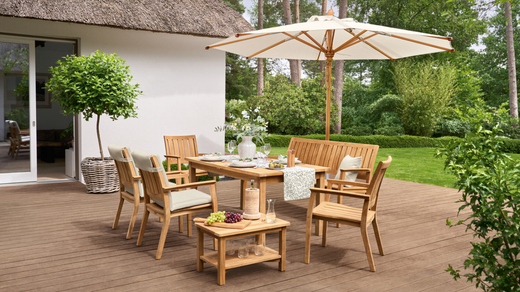 A wooden patio dining set with chairs and a bench sits on a deck beside a white house, under a large cream-colored umbrella. The table is set for a meal, surrounded by greenery and trees.
