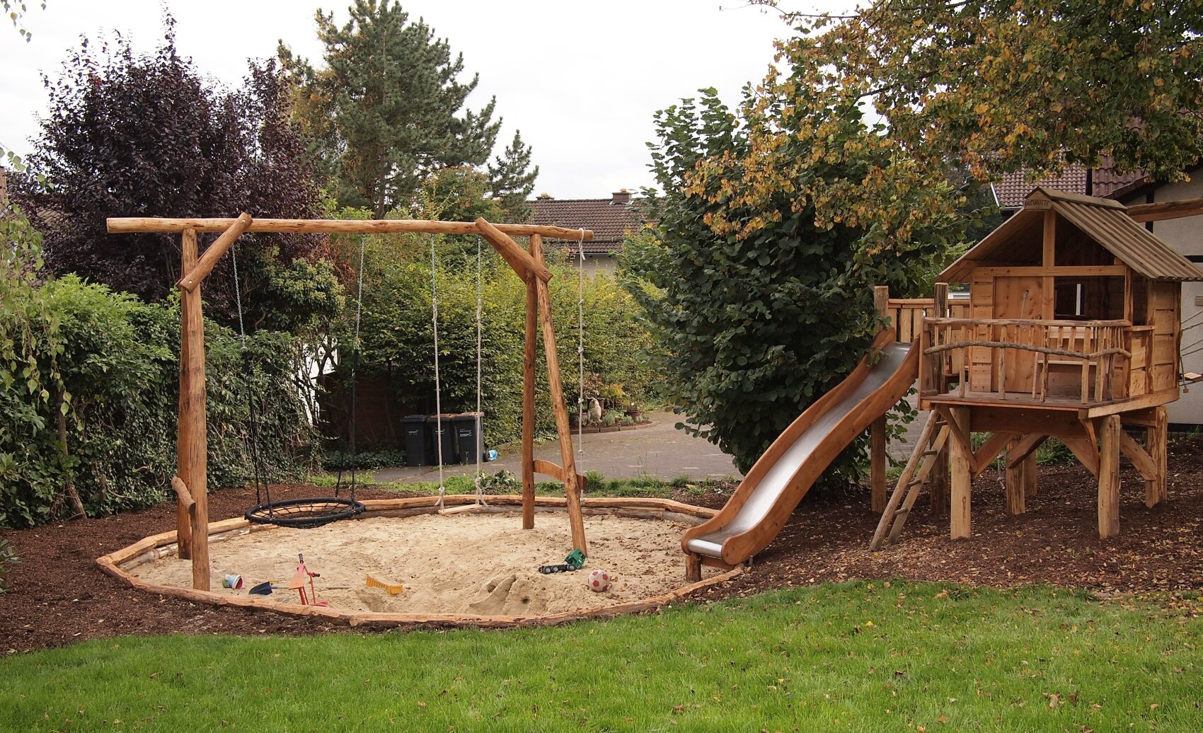 A backyard playground with a wooden swing, a sandbox, a slide, and a wooden playhouse, surrounded by trees and grass.
