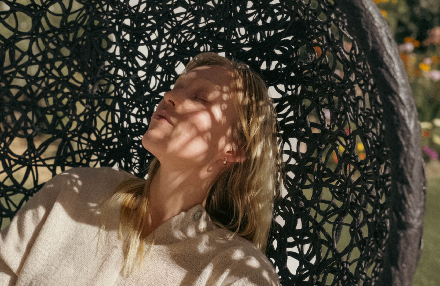 Eine Frau mit blondem Haar lehnt sich in einem geflochtenen Stuhl im Freien zurück, die Augen geschlossen, während Sonnenlicht und Schatten Muster auf ihrem Gesicht und ihrem leichten Pullover erzeugen. Grünzeug und Blumen sind im Hintergrund verschwommen.