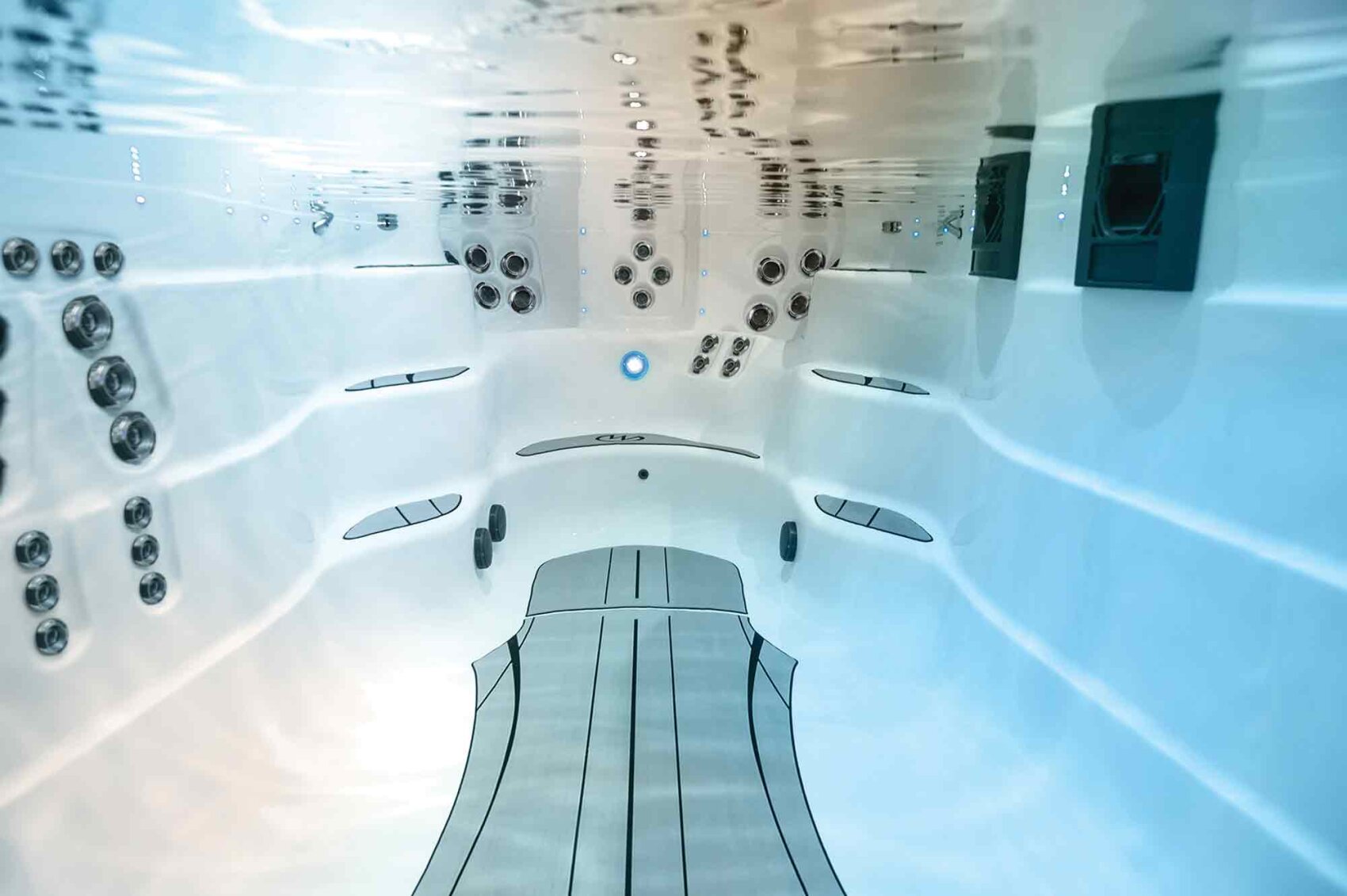 Underwater view inside an empty white swim spa or pool, showing built-in seating, multiple water jets, and overhead lighting, with a clear, reflective water surface above.