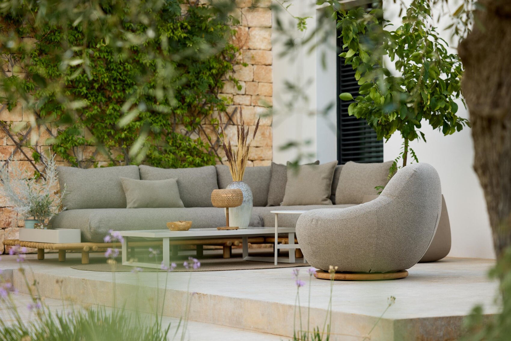 Terrasse im Freien mit einem modernen grauen Sektionssofa, einem runden Sessel und niedrigen Couchtischen auf einer Steinterrasse, umgeben von Grün und Pflanzen, neben einer weißen Ziegelwand.