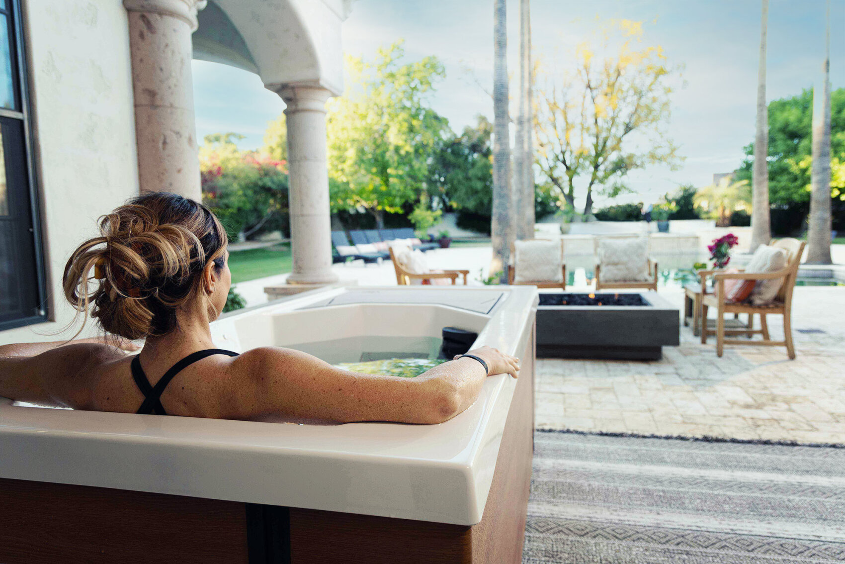 A woman relaxes in an outdoor hot tub, facing a patio with chairs and a fire pit, surrounded by trees and greenery on a sunny day.