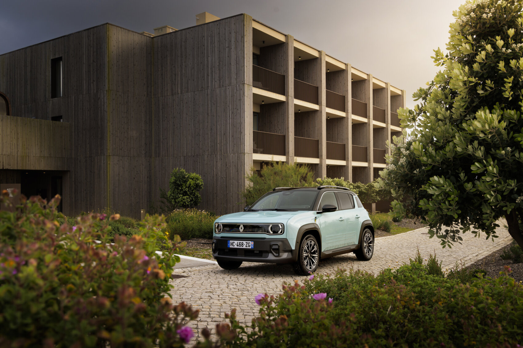 Ein hellblauer elektrischer Geländewagen parkt auf einer kopfsteingepflasterten Einfahrt vor einem modernen, mehrstöckigen Gebäude mit Holzwänden, umgeben von grünen Sträuchern und blühenden Pflanzen unter einem bewölkten Himmel.
