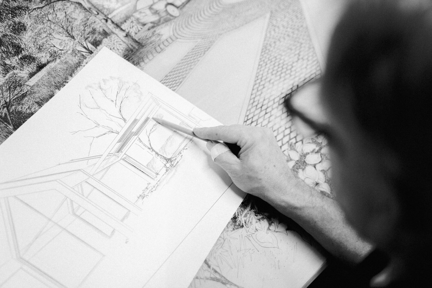 Eine Person, die eine Brille trägt, zeichnet architektonische Zeichnungen eines Hauses auf Papier, umgeben von anderen detaillierten Zeichnungen und Plänen.