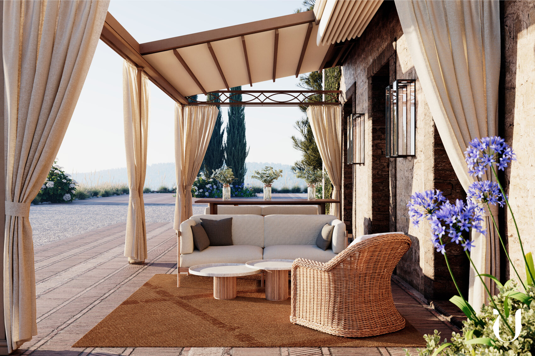 A cozy outdoor patio with cream-colored curtains, a wicker armchair, a cushioned sofa, two round tables, and potted purple flowers on a brown rug, set against a rustic building with trees and mountains in the background.