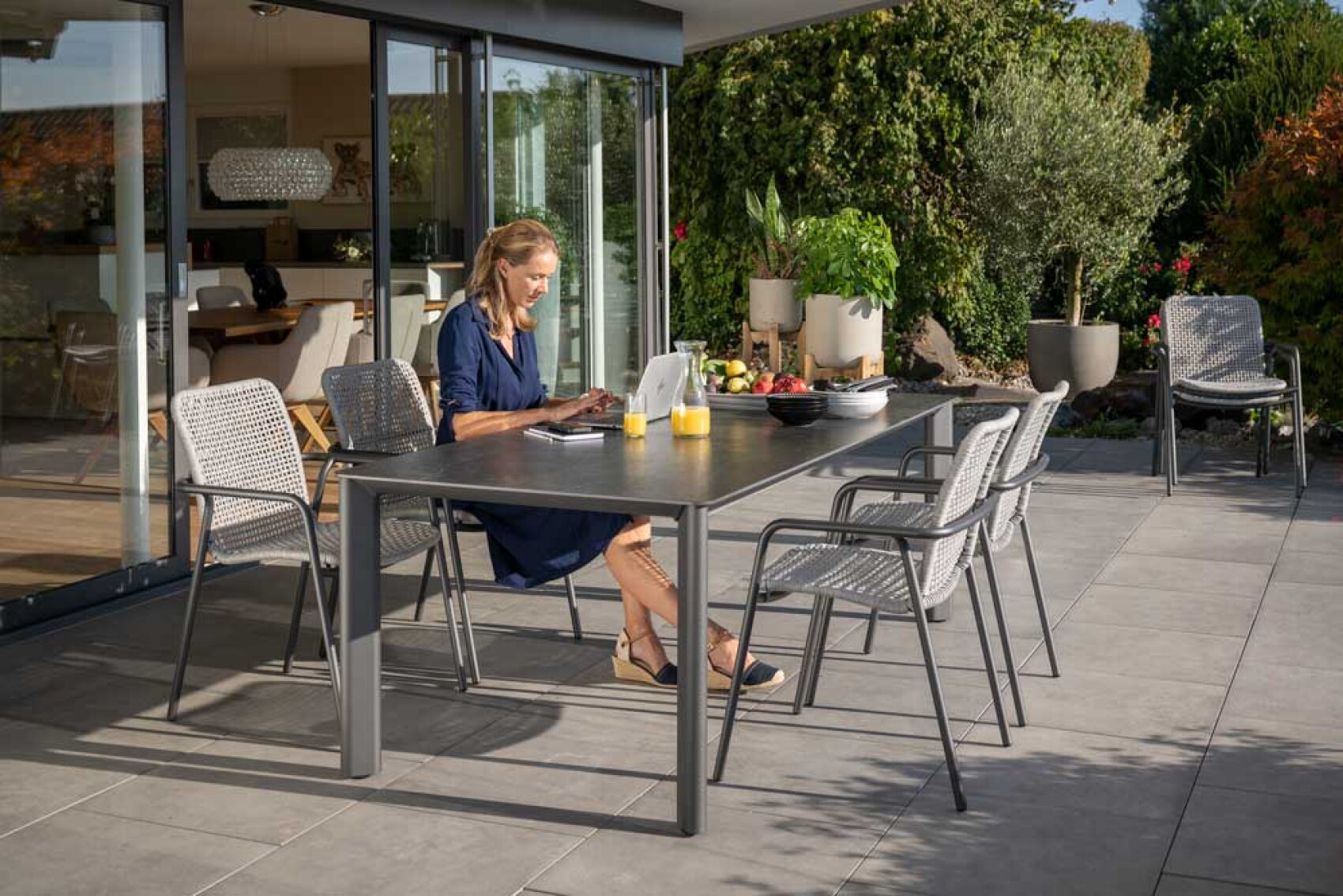 Eine Frau sitzt an einem Tisch auf einer sonnigen Terrasse und arbeitet an einem Laptop. Auf dem Tisch stehen Getränke, Obst und eine Schale, um die herum leere Stühle stehen. Glastüren öffnen sich zu einer modernen Küche, und der Innenhof ist von Grün umgeben.