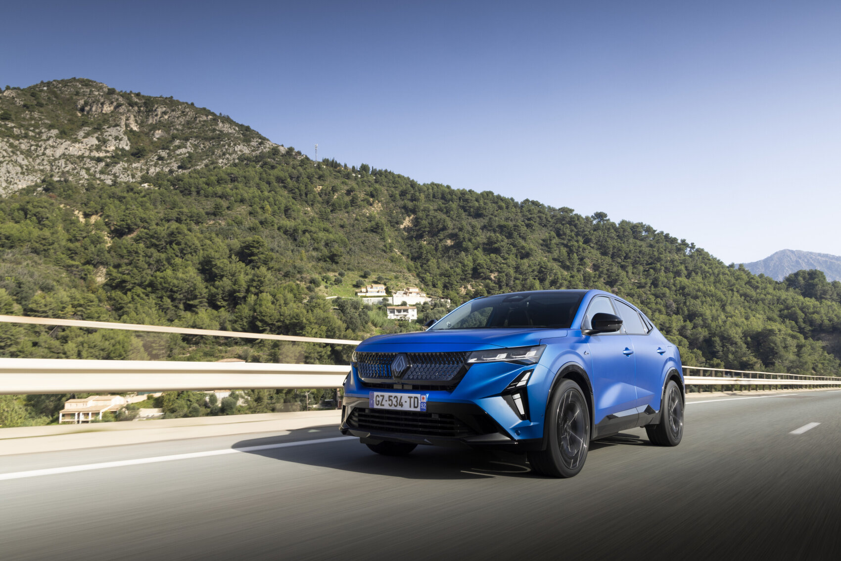 Ein blauer Geländewagen fährt auf einer sonnigen Landstraße, umgeben von grünen Hügeln und Bergen, mit einem klaren blauen Himmel im Hintergrund.