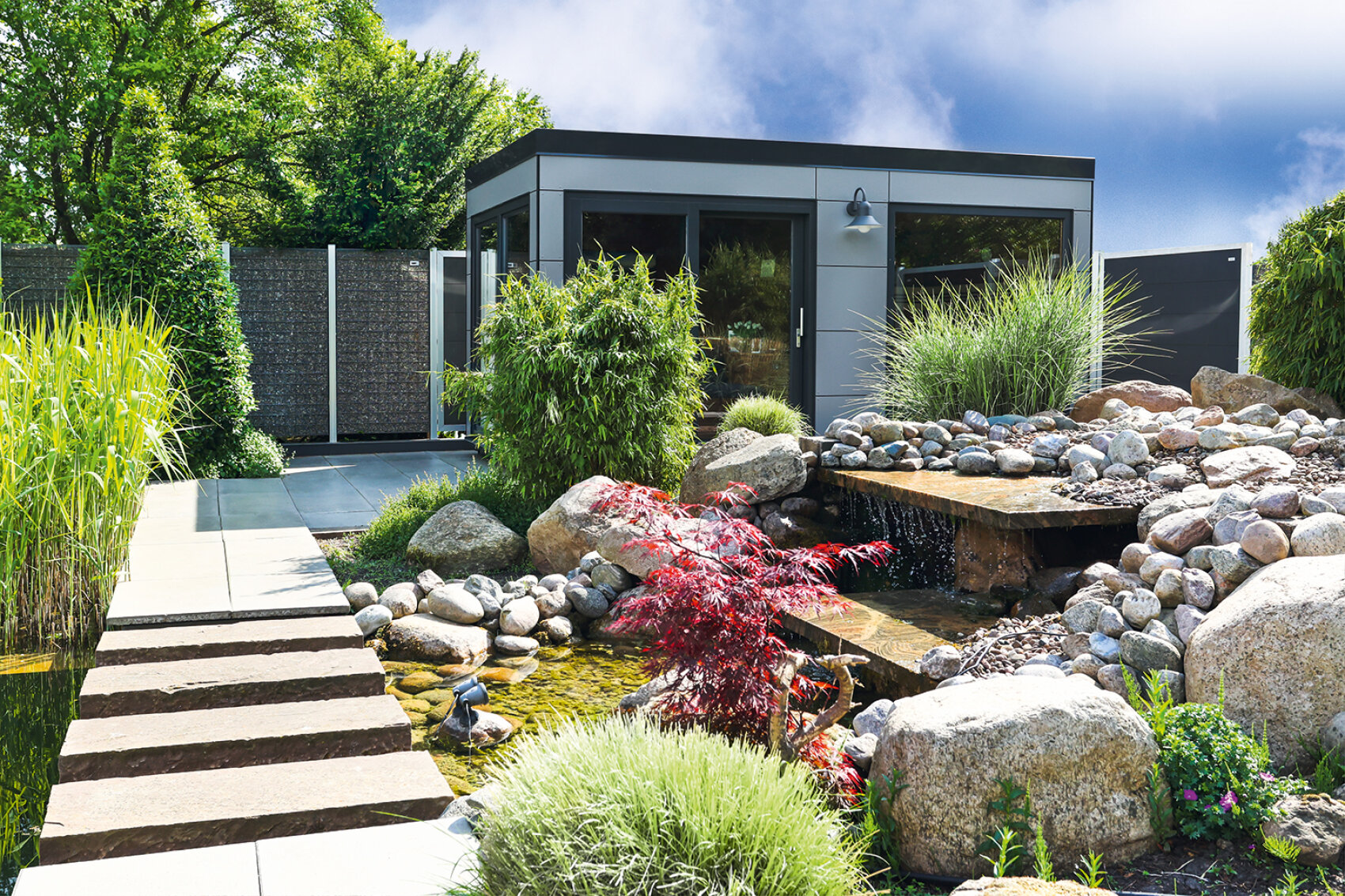 A modern garden with a sleek, gray garden shed, a small pond with rocks, a wooden stepping path, lush green plants, and a red-leaved shrub, all under a partly cloudy sky.