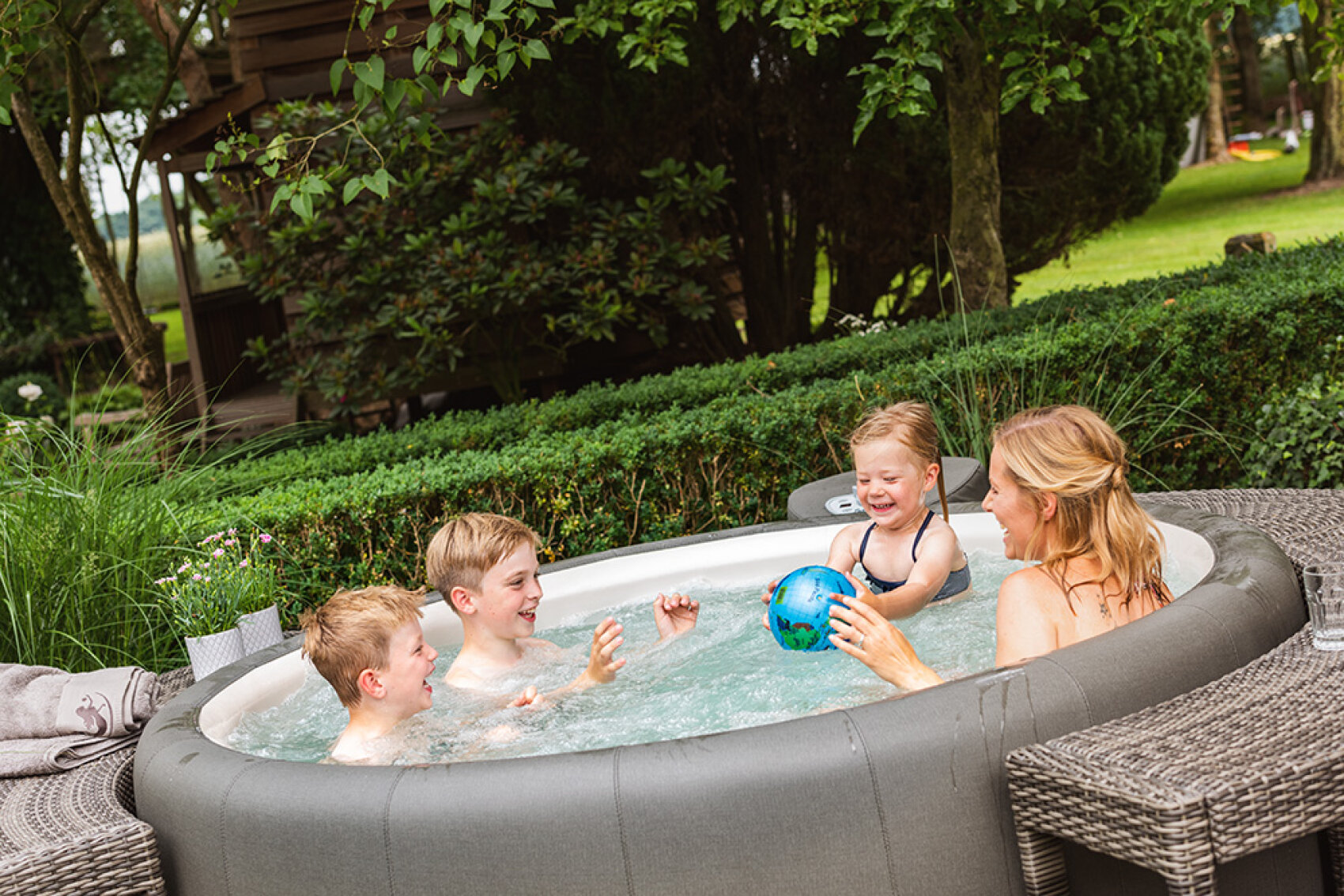 A woman and three young children play with a blue ball in an outdoor inflatable hot tub, surrounded by greenery and garden furniture on a sunny day.