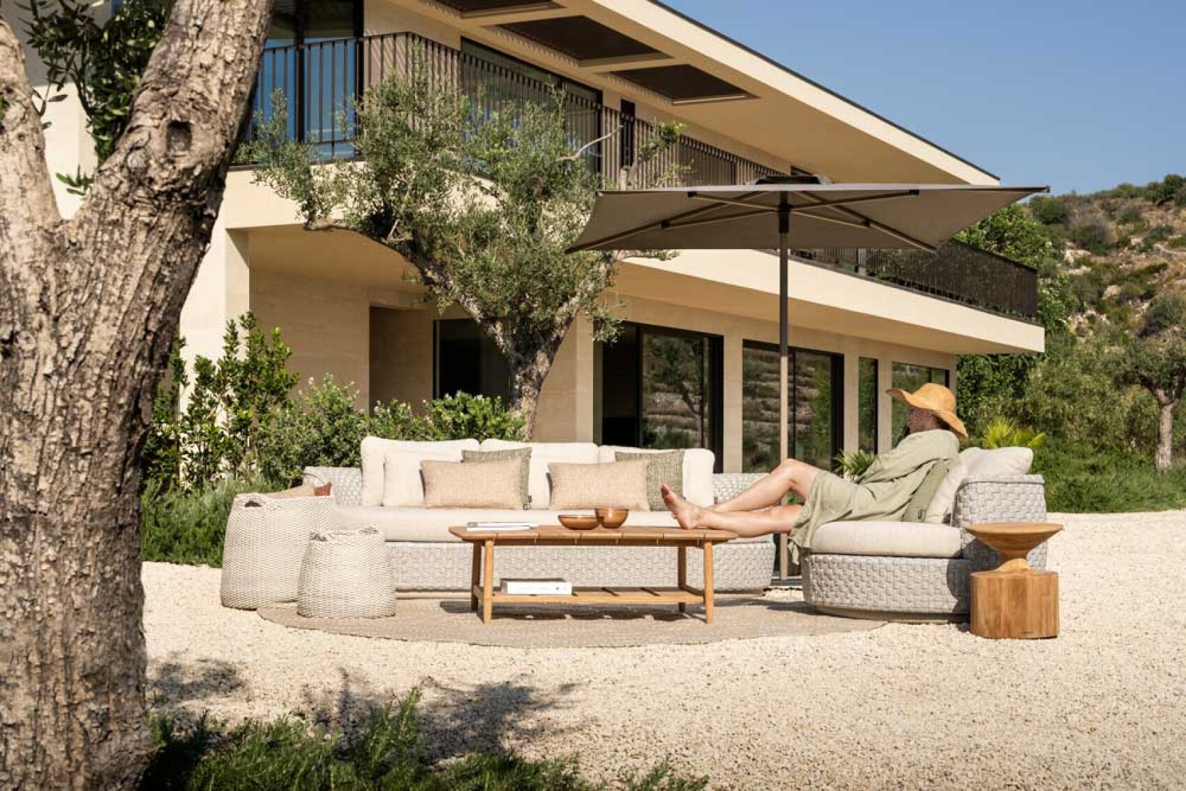 Eine Person mit Sonnenhut entspannt sich auf einem modernen Outdoor-Sofa unter einem großen Sonnenschirm in einem angelegten Garten mit Bäumen vor einem modernen Haus mit großen Fenstern und einem Balkon.