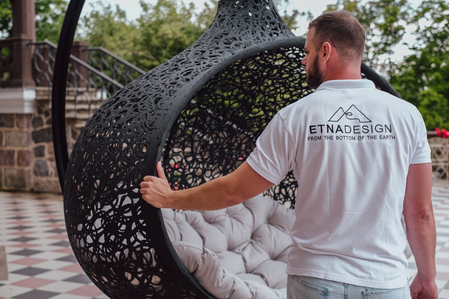 Ein Mann in einem weißen "ETNADESIGN"-Hemd steht neben einem modernen, schwarzen, geflochtenen Outdoor-Hängesessel mit einem weißen Kissen auf einer karierten Terrasse. Im Hintergrund sind Bäume und Steinmauern zu sehen.
