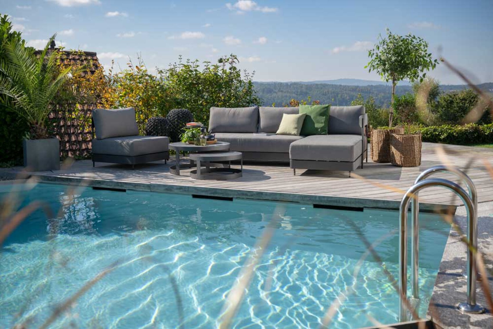 Eine moderne Außenterrasse mit grauen Polstermöbeln liegt neben einem Swimmingpool unter blauem Himmel, umgeben von Grünpflanzen und Kübelpflanzen.
