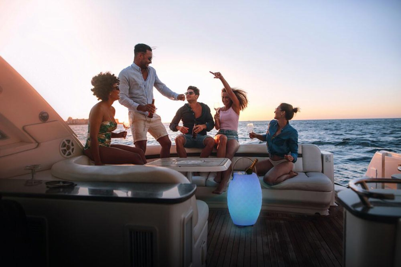 Fünf Personen entspannen sich bei Sonnenuntergang auf einer Yacht und genießen einen Drink, lachen und reden miteinander. Im Hintergrund sind das Meer und der Horizont zu sehen, und auf dem Deck befindet sich eine leuchtende dekorative Lampe.