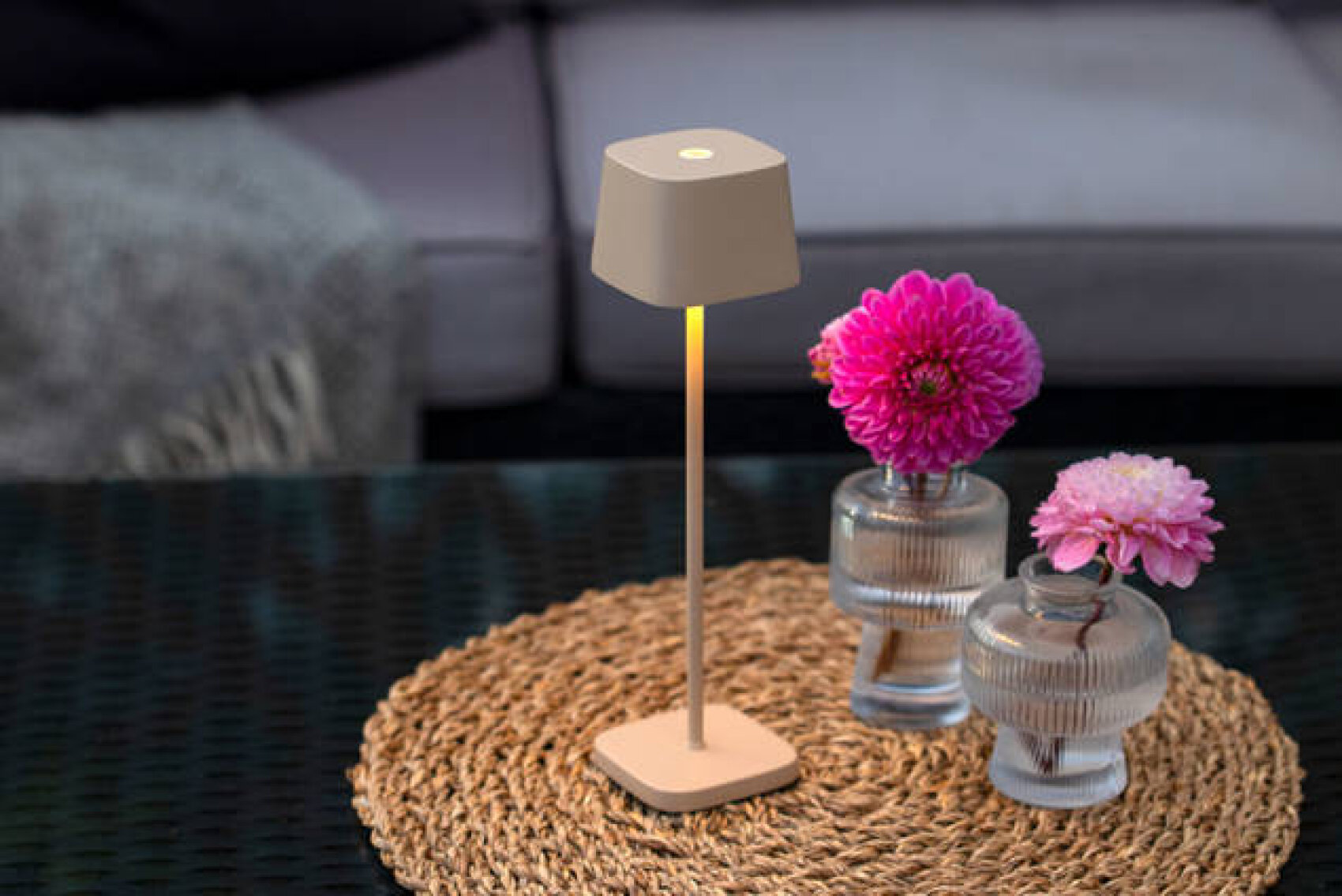 Eine moderne beigefarbene Tischlampe und zwei durchsichtige Vasen mit rosafarbenen Blumen stehen auf einem gewebten Tischset auf einem dunklen Glastisch, mit einem grauen Sofa und einer Decke im Hintergrund.