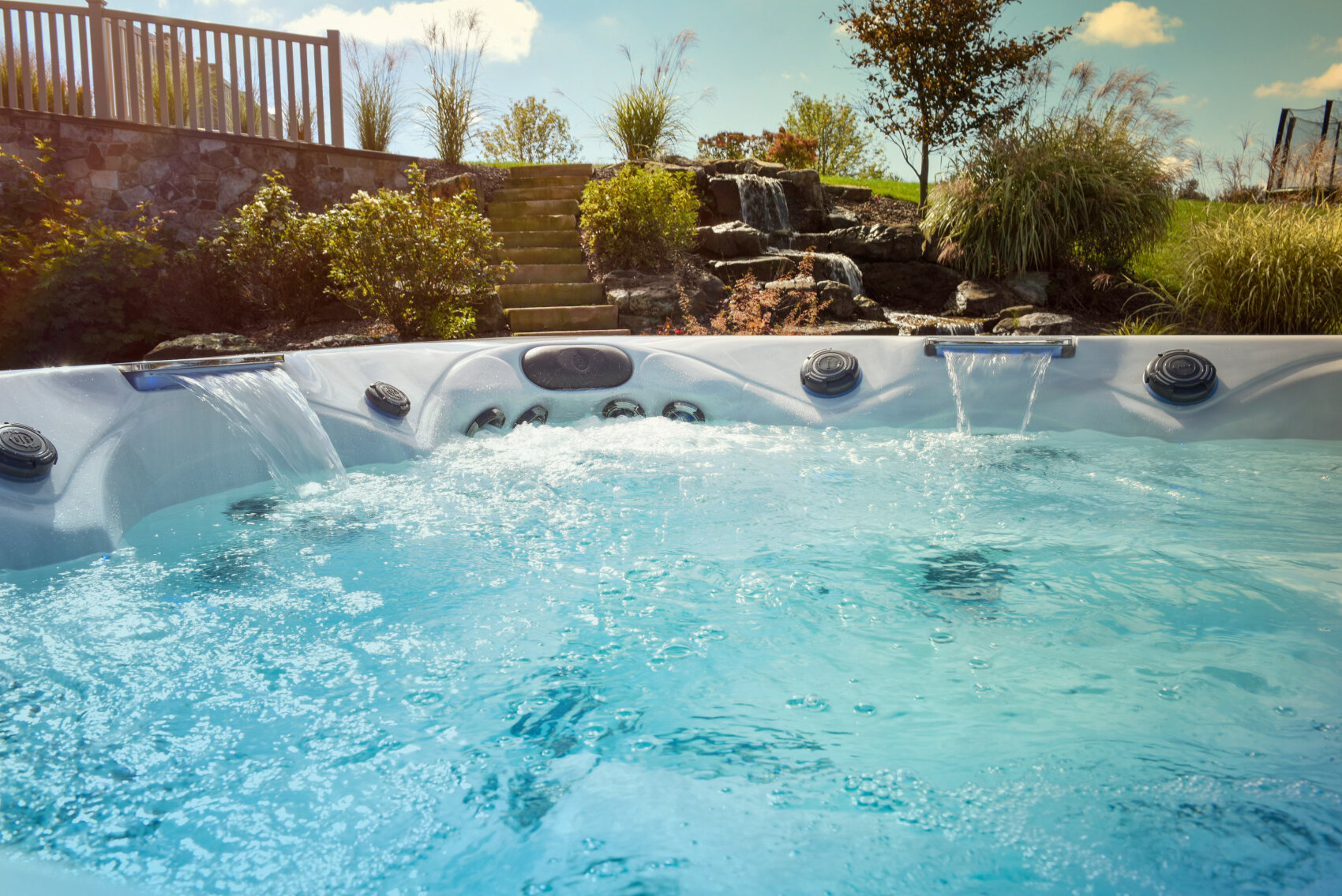 A bubbling hot tub with clear water sits outdoors, surrounded by lush landscaping, tall grasses, stone steps, and a waterfall feature under a blue sky with some clouds.