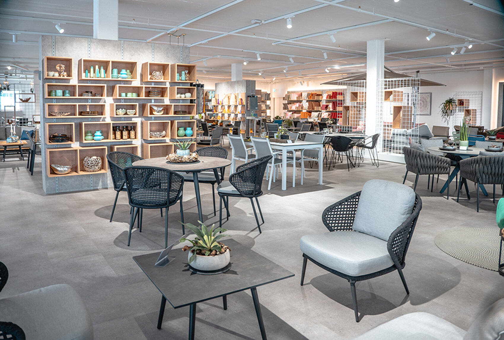 Ein moderner Möbel-Showroom mit Tischen, Stühlen und Regalen, in denen Dekorationsartikel ausgestellt sind. Der Raum ist gut beleuchtet und verfügt über eine klare, offene Aufteilung und eine Vielzahl von Sitzmöglichkeiten.