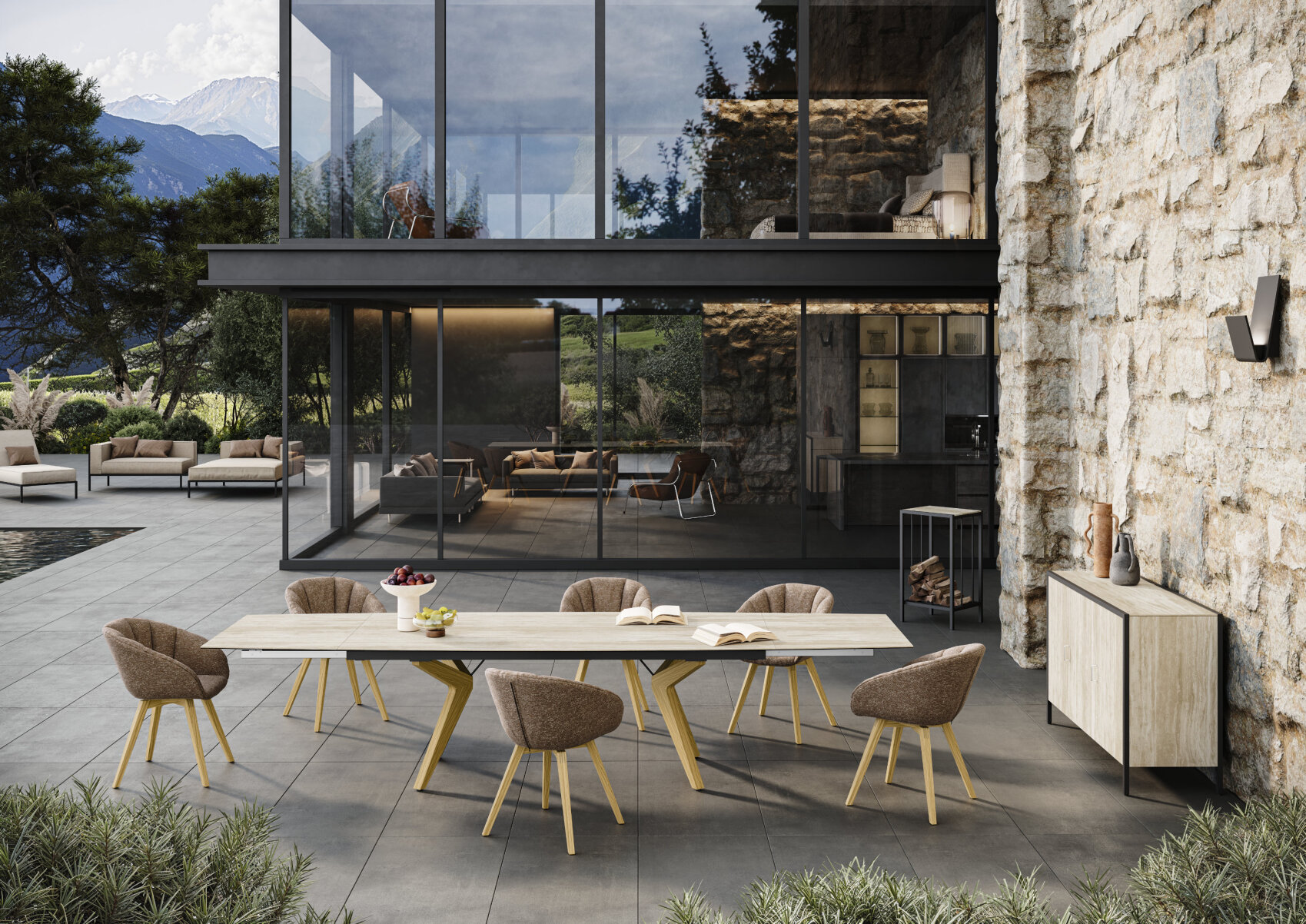Modern outdoor patio with a long dining table and six brown chairs on a tiled terrace. There is a stone wall, floor-to-ceiling windows, and a pool, with trees and mountains visible in the background.