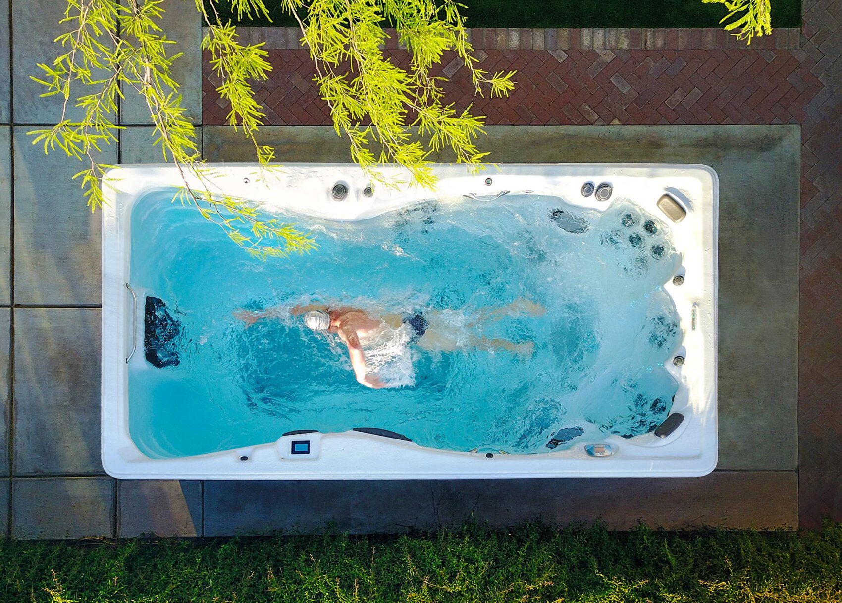 A person wearing a swim cap swims laps in a swim spa pool outdoors, surrounded by greenery and brick pavement, as seen from above.