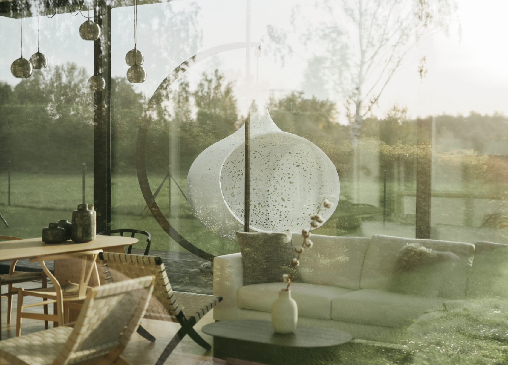 Ein gemütliches, modernes Wohnzimmer mit weißem Sofa, Holzmöbeln und einem einzigartigen Hängesessel, der sich in großen Glasfenstern mit Blick auf eine grüne Landschaft bei Sonnenuntergang spiegelt.