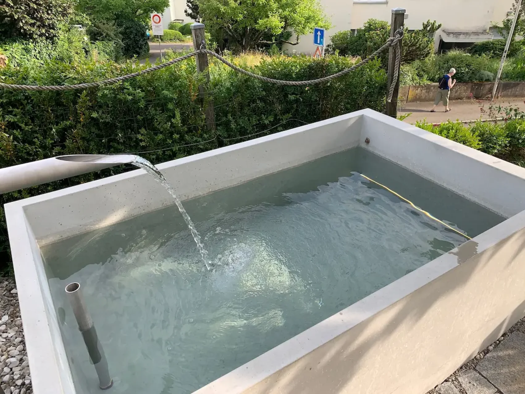 Ein rechteckiger Wassertrog aus Beton ist mit klarem Wasser gefüllt, in das ein Metallrohr Wasser einleitet. Im Hintergrund sind grüne Sträucher und ein Wanderweg mit einer Person zu sehen.