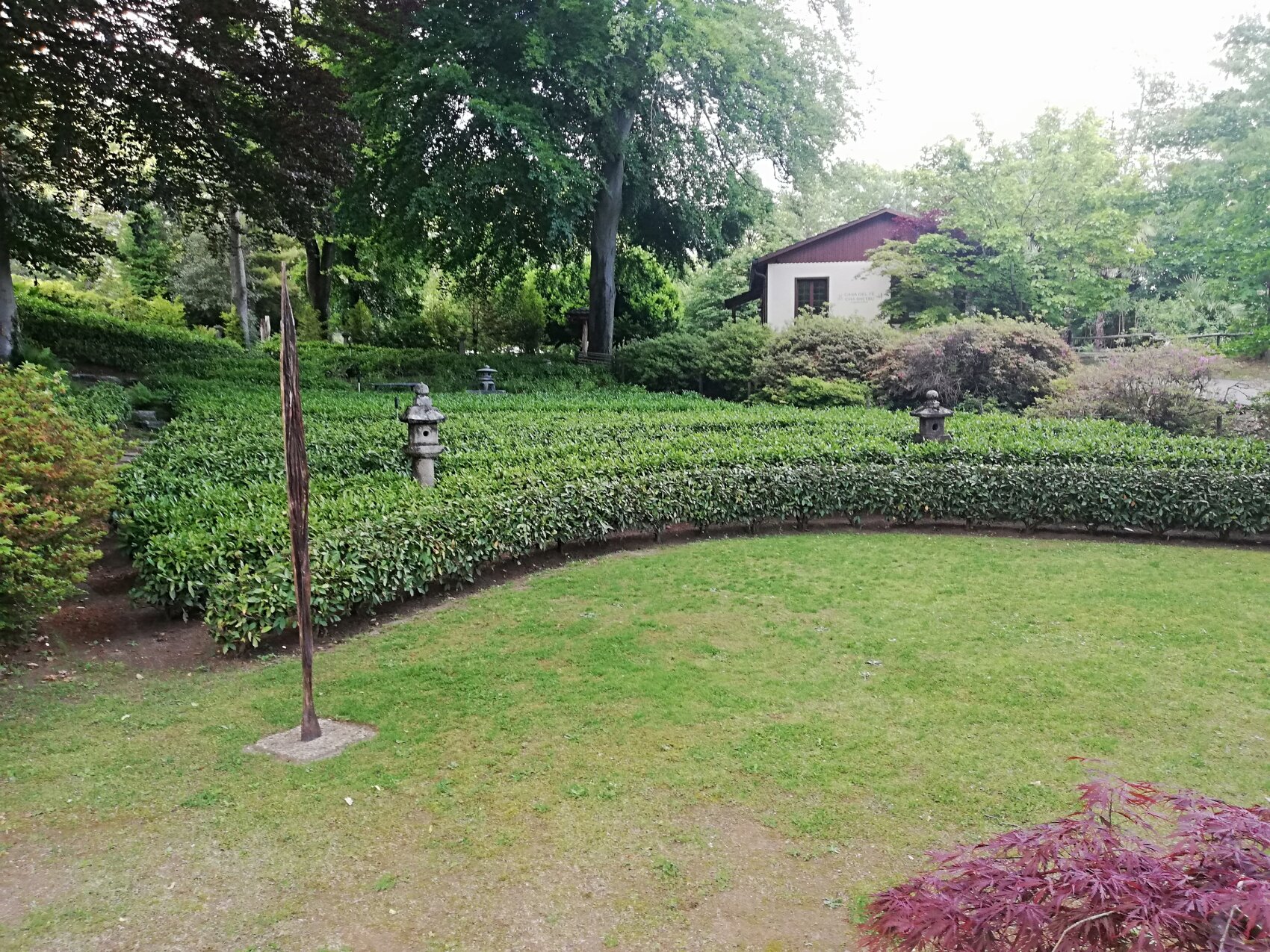 Ein üppiger Garten mit ordentlich geschnittenen Hecken, grünem Gras, Steinlaternen und einem kleinen weißen Gebäude im Hintergrund, umgeben von hohen Bäumen und belaubten Sträuchern.