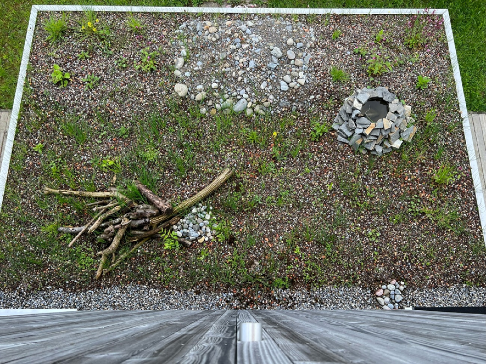 Ein rechteckiger Fleck aus Kies und spärlichem Gras enthält verstreute Steine, einen Haufen Äste und einen kreisförmigen Ring aus Steinen, der eine kleine Grube umgibt (von oben betrachtet). Der untere Rand wird von einem Holzdeck begrenzt.