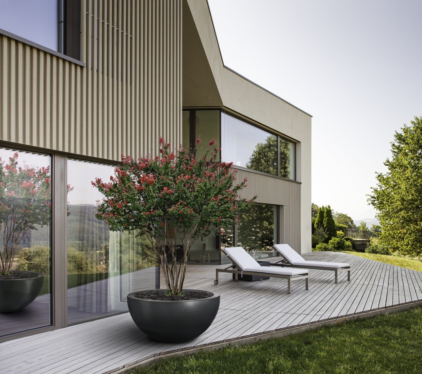 Ein modernes Haus mit großen Fenstern und einer Holzterrasse mit zwei weißen Liegestühlen und einer großen Topfpflanze mit rosa Blüten, umgeben von grünem Gras und Bäumen an einem sonnigen Tag.